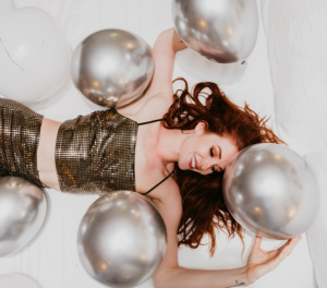 red headed girl laying in hotel bed with gold glittery outfit on surrounded by balloons. the girl is smiling.