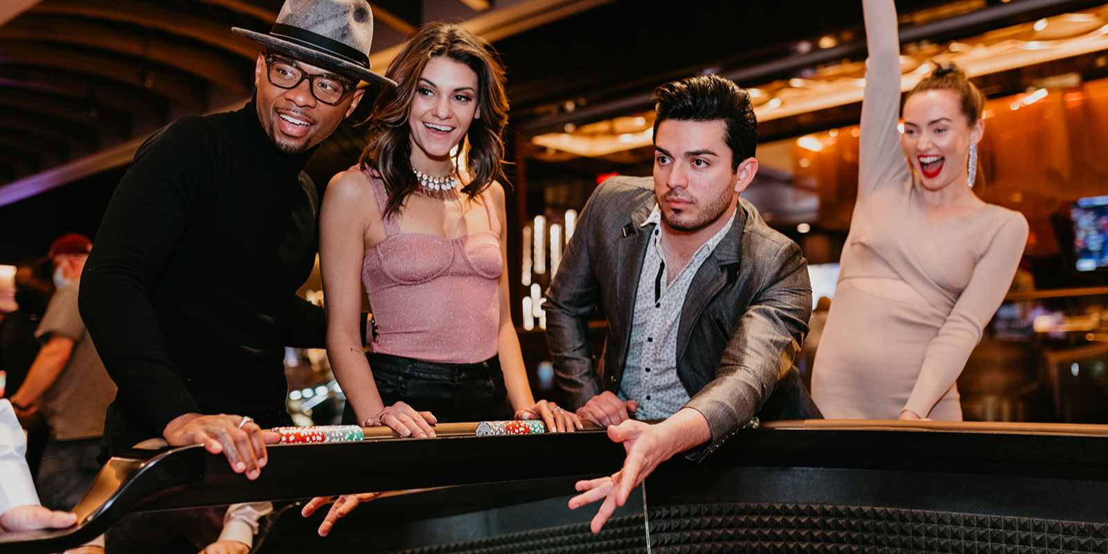 Four people standing around a craps table. One guy is throwing his dice to play while his 3 friends watch. One girl is celebrating with her right arm raised to the roof. Night time casino vibes.