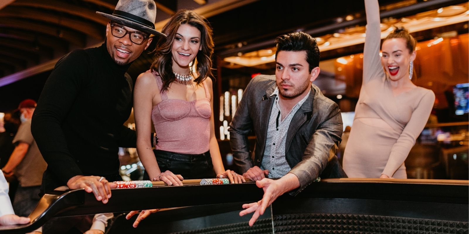 Four people standing around a craps table. One guy is throwing his dice to play while his 3 friends watch. One girl is celebrating with her right arm raised to the roof. Night time casino vibes.