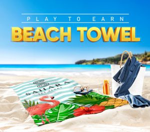 visual that promotes a beach towel giveaway. It says Play To Earn Beach Towel and shows the beach towel on the beach near the ocean with a beach bag sandals and a water bottle