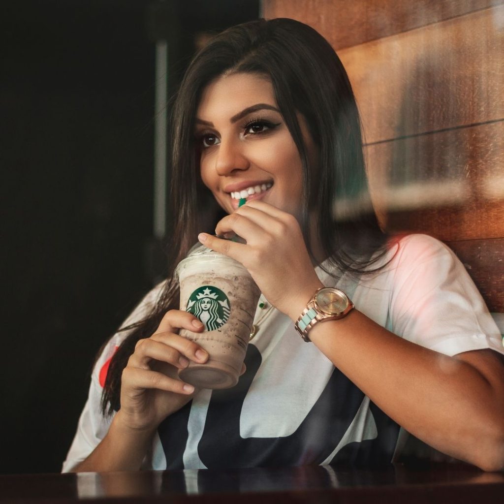 a young twenty something girl with brown hair smiling as she sips on her Starbucks frappaccino
