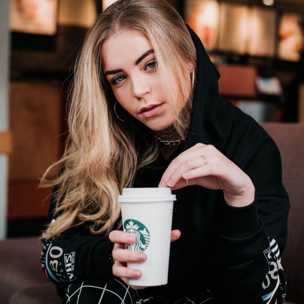 A young looking girl with blonde hair and blue eyes holding a Starbucks cup