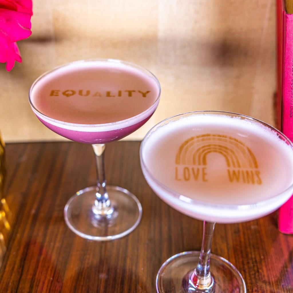 Two cocktails sitting on a table with text floating on the top of the drinks. One says Love Wins and shows a rainbow and the other says equality