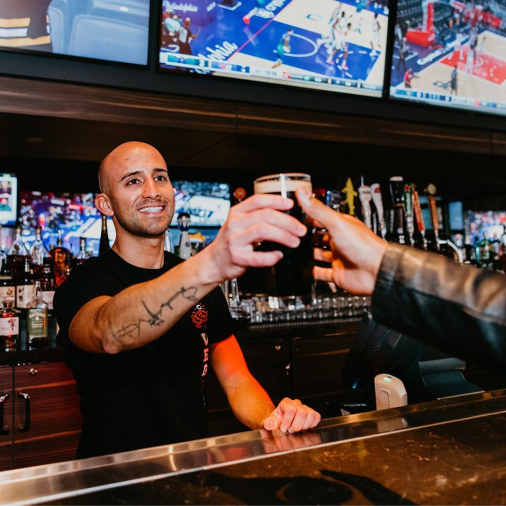 Chickie's & Pete's bartender handing a guest a beer. The picture shows the bartender smiling and standing in front of several tv screens showing sports games