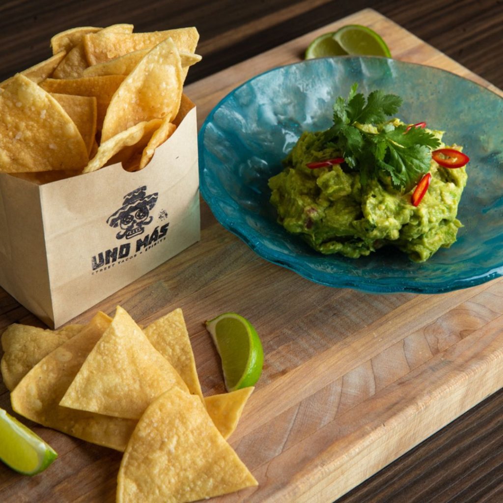 Uno Mas guacamole in a blue bowl with chips on a wooden cutting board