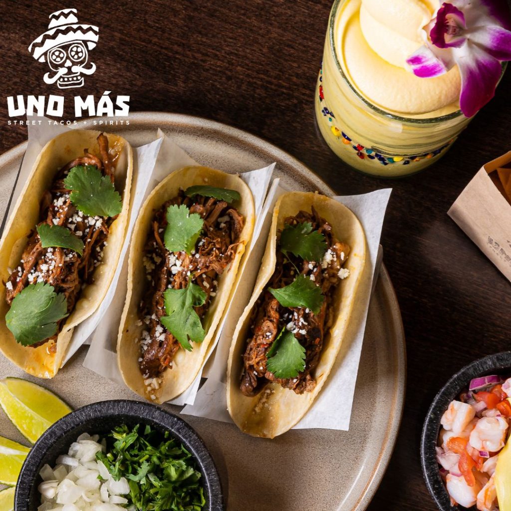 Uno Mas street tacos on a plate next to a blended yellow mnargarita and chips with limes, onion and cilantro