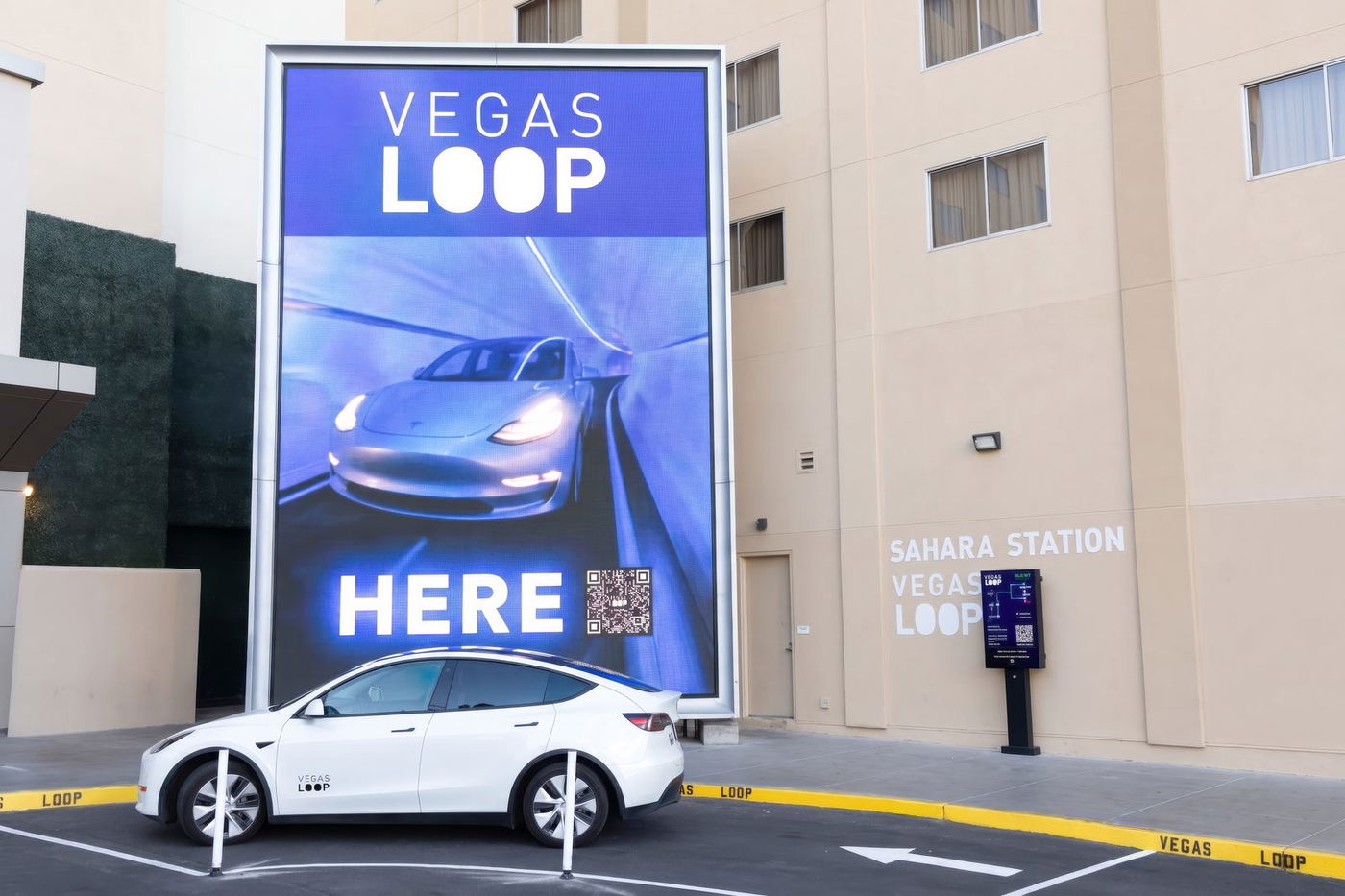 The Vegas Loop station at SAHARA Las Vegas, with a tall illuminated Vegas Loop marquee screen, SAHARA STATION signage on the building, a ticket kiosk, and a white Tesla waiting at the curb.